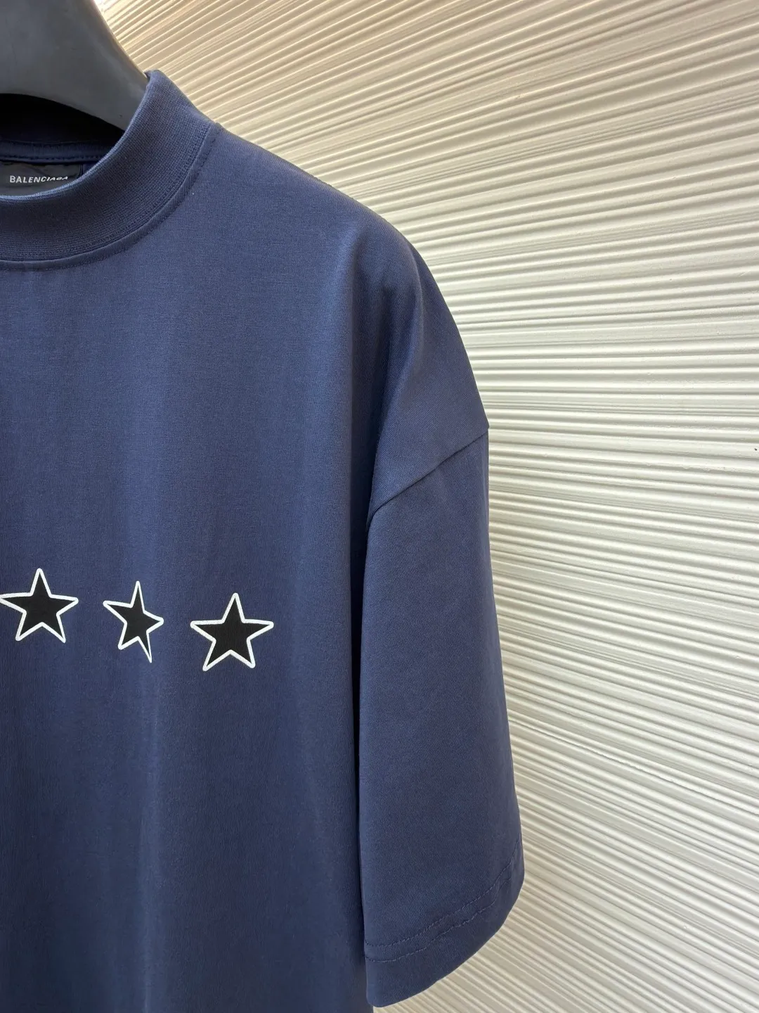 Balenciaga star embroidered short sleeves [comes with marker] ●Item number: 1682275-A20 ●Material: combed cotton fabric ●Technology: embroidery printing ●Color: blue ●Version: loose fit ●Weight: about 355 grams ●Size: XS-S-M-L ●Description: This navy blue short-sleeved T-shirt is made of high-density piqué mesh cotton fabric. The fabric has been custom-woven and dyed. The navy blue tone is uniform and not easy to fade. The breathable structure formed by the piqué texture makes the wearer more refreshing. At the same time, the fabric has excellent stiffness and can support the loose shoulder silhouette. The five-star pattern on the chest adopts double-track offset printing + inlay embroidery process. First, the outline of the stars is outlined with high-temperature embossing using matte silicone material. The thickness of the silicone layer is controlled at 0.3mm to ensure softness and no irritation to the skin. White embroidery thread is then used to fill in inlay along the edge of the silicone to give the star pattern a three-dimensional relief texture. The stitch density of each star reaches 12 stitches/cm, and the pattern alignment error does not exceed 0.1cm. The neckline is designed with a small stand-up collar, which is woven with ribs of the same fabric. It is not easy to loose after high-temperature pre-shrinking treatment. The side seams are spliced ​​with a seamless buried clip process to reduce friction when wearing. The overall process takes into account the texture and wearing comfort. The overall wiring is straight and delicate, the fabric is comfortable and soft, and the color is consistent without color difference. It can be matched with the same style to create a fashionable look and control various occasions. Customized accessories, complete with three standards, same style for men and women!