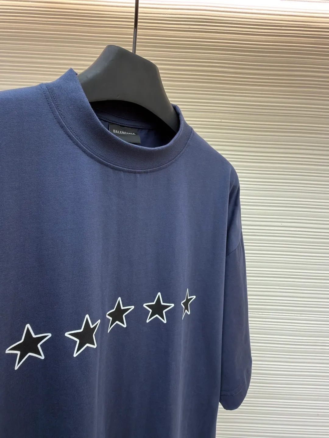 Balenciaga star embroidered short sleeves [comes with marker] ●Item number: 1682275-A20 ●Material: combed cotton fabric ●Technology: embroidery printing ●Color: blue ●Version: loose fit ●Weight: about 355 grams ●Size: XS-S-M-L ●Description: This navy blue short-sleeved T-shirt is made of high-density piqué mesh cotton fabric. The fabric has been custom-woven and dyed. The navy blue tone is uniform and not easy to fade. The breathable structure formed by the piqué texture makes the wearer more refreshing. At the same time, the fabric has excellent stiffness and can support the loose shoulder silhouette. The five-star pattern on the chest adopts double-track offset printing + inlay embroidery process. First, the outline of the stars is outlined with high-temperature embossing using matte silicone material. The thickness of the silicone layer is controlled at 0.3mm to ensure softness and no irritation to the skin. White embroidery thread is then used to fill in inlay along the edge of the silicone to give the star pattern a three-dimensional relief texture. The stitch density of each star reaches 12 stitches/cm, and the pattern alignment error does not exceed 0.1cm. The neckline is designed with a small stand-up collar, which is woven with ribs of the same fabric. It is not easy to loose after high-temperature pre-shrinking treatment. The side seams are spliced ​​with a seamless buried clip process to reduce friction when wearing. The overall process takes into account the texture and wearing comfort. The overall wiring is straight and delicate, the fabric is comfortable and soft, and the color is consistent without color difference. It can be matched with the same style to create a fashionable look and control various occasions. Customized accessories, complete with three standards, same style for men and women!