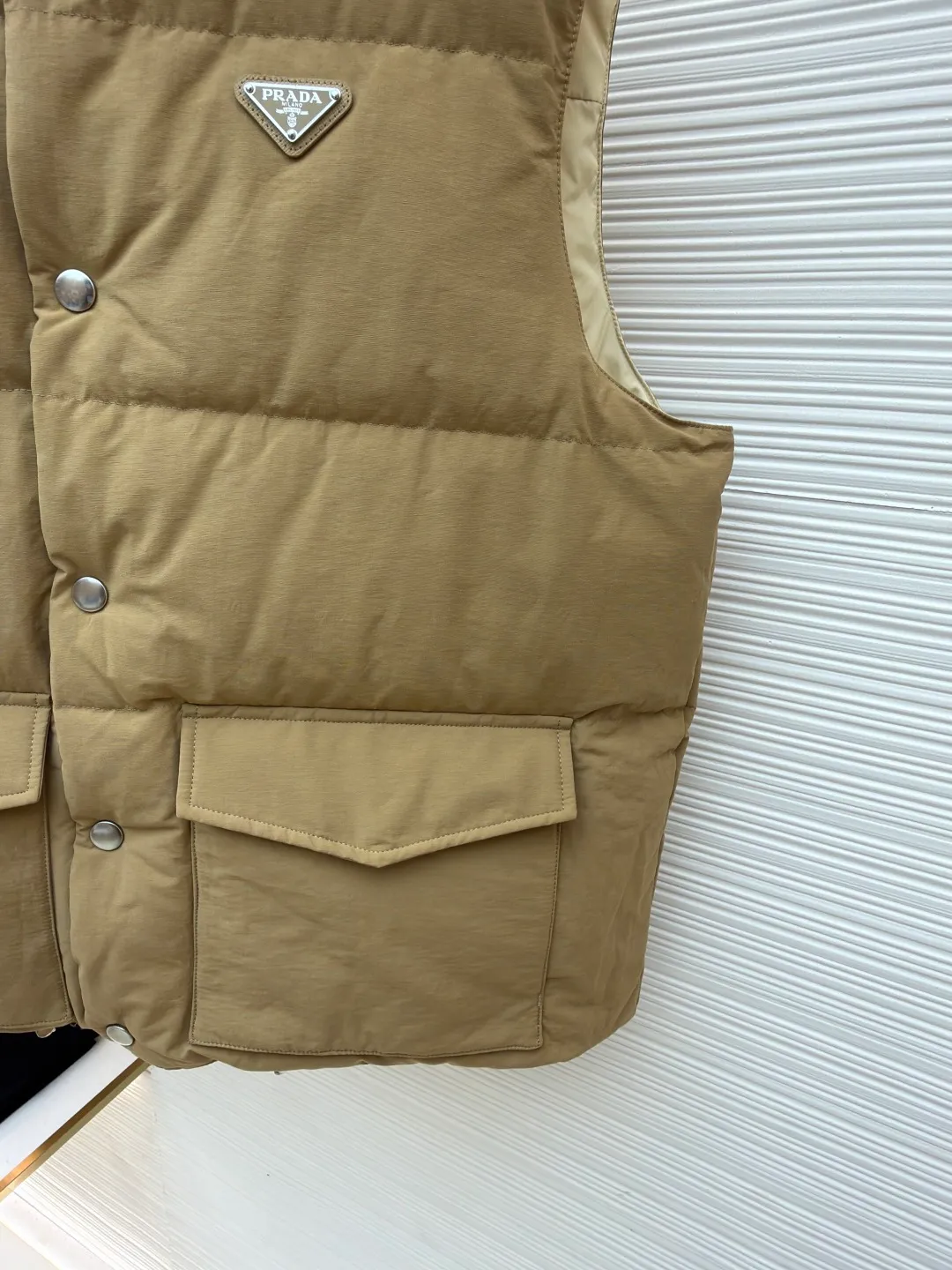 Prada stand collar badge down vest ●Original version: purchased and developed by 18756 ●Item number: 16801377-C30 ●Fabric: white duck down fabric ●Technology: mechanical down filling ●Color: khaki ●Version: Fitted version ●Weight: about 700 grams ●Size: S-M-L-XL ●Description: This vest is made of high-quality water-repellent fabric, and the khaki tone shows the texture. Using three-dimensional quilting technology, the down is evenly divided into full grid-shaped down-filled areas, which not only ensures warmth but also makes the silhouette crisp and three-dimensional. The neckline is designed with a stand-up collar and is matched with a metal snap placket, which is exquisite in texture and easy to put on and take off.  The front chest is decorated with the brand's iconic metal triangle logo, which is highly recognizable; the large patch pockets on both sides are three-dimensionally cut, and the bag mouth is in an inverted trapezoid shape, combining practicality and design. There is a built-in drawstring at the hem, and the tightness can be adjusted freely to suit different wear needs. From fabric selection to detail sewing, every process reflects the brand's strict control of craftsmanship, exquisitely integrating functionality and fashion sense. The overall wiring is straight and delicate, the fabric is comfortable and soft, and the color is consistent without color difference. It can be matched with the same style to create a fashionable look and control various occasions. Customized accessories, complete with three standards, same style for men and women!