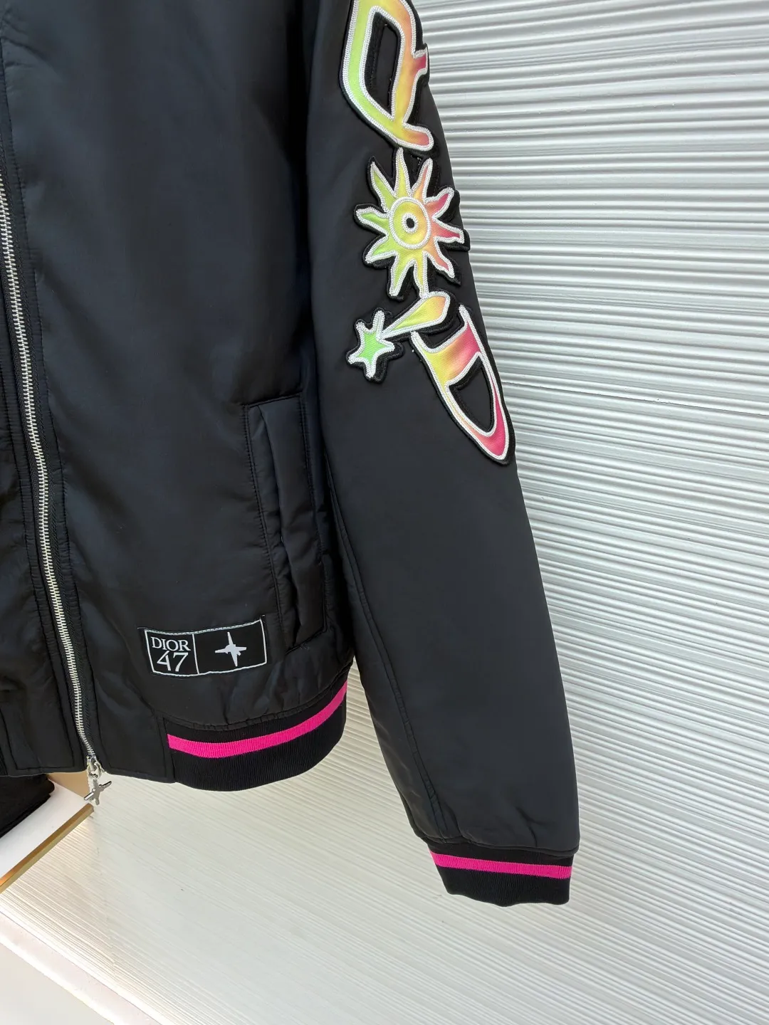 Dior Hamilton joint baseball uniform ●Original version: 32000 purchased and developed ●Item number: 1681336-B90 ●Fabric: nylon fabric ●Technology: printing production ●Color: black ●Version: fitted version ●Weight: about 830 grams ●Size: 48-50-52-54 ●Description: Made of high-quality matte nylon fabric, the texture is stiff and windproof. It has been treated with a special coating to improve wear resistance and texture, and provide good shape support for the jacket. The colorful letter pattern on the sleeves uses a multi-layer offset printing process, with natural color gradients and strong adhesion; the "DIOR 47" logo on the chest is precision embroidered, with fine stitching and high integration with the fabric, highlighting the brand's craftsmanship. Made from a classic baseball uniform cut, the silhouette is sleek and stylish. Double thread reinforcement is used during sewing, the seams are smooth and snug, the stitch spacing is even, and the lines are straight, reflecting the rigorous garment making process. The zipper is a brand-customized metal zipper with brand details on the zipper puller, which is smooth and durable; the ribbed collar, cuffs and hem are made of high-elastic knitting technology, which has good elasticity, is comfortable to wear and can effectively prevent wind. The pockets on both sides are designed as hidden pockets, which are practical while maintaining overall beauty; the pink trim on the body contrasts sharply with the main black tone, and the craftsmanship details reflect the brand's high-end production standards.