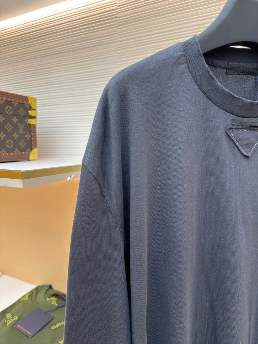 Prada Pencil Gray Long Sleeve ●Original version: Purchased and developed by 8934 ●Item number: 1680720-A05 ●Fabric: combed plain weave fabric ●Technology: Laser burning ●Version: fitted version ●Weight: about 240 grams ●Size: S-M-L-XL ●Description: Custom-woven 50S 190G combed plain weave fabric, non-market general gray fabric, needs to be fully customized from yarn ratio and loom parameters. Short fiber impurities are removed through the combing process to ensure that the yarn is longer and more uniform, and the fabric surface is smooth and hairless.  Specially dyed pencil gray, the color is adjusted individually instead of using a standard color card. Multiple proofing is required to match the low saturation gray tone specified by Prada. Chest triangle laser burnt embroidery shaping: Laser burnt: high-precision laser engraving, carbonizing the fabric surface to form a gradient etching texture, and the edges need to be manually adjusted to avoid scorch marks.  Composite embroidery: superimpose embroidery on the basis of burnt flowers. The stitch density needs to be aligned with the burnt flowers pattern to prevent fabric deformation.  Three-dimensional shaping: Use high-temperature pressing or resin coating to maintain the relief of the triangular structure and avoid collapse after washing. The overall wiring is straight and delicate, the fabric is comfortable and soft, and the color is consistent without color difference. It can be matched with the same style to create a fashionable look and control various occasions. Customized accessories, complete with three standards, same style for men and women!