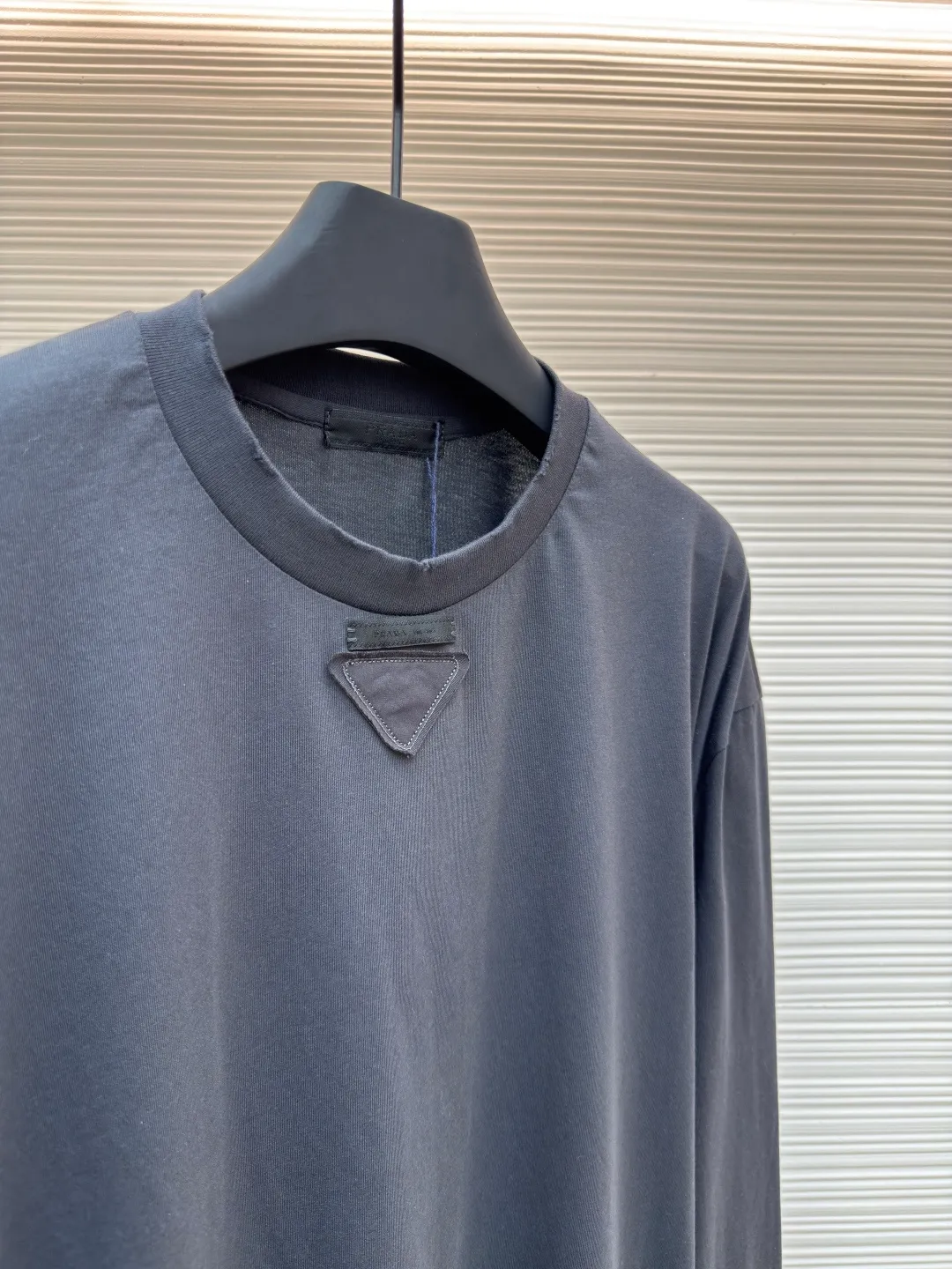 Prada Pencil Gray Long Sleeve ●Original version: Purchased and developed by 8934 ●Item number: 1680720-A05 ●Fabric: combed plain weave fabric ●Technology: Laser burning ●Version: fitted version ●Weight: about 240 grams ●Size: S-M-L-XL ●Description: Custom-woven 50S 190G combed plain weave fabric, non-market general gray fabric, needs to be fully customized from yarn ratio and loom parameters. Short fiber impurities are removed through the combing process to ensure that the yarn is longer and more uniform, and the fabric surface is smooth and hairless.  Specially dyed pencil gray, the color is adjusted individually instead of using a standard color card. Multiple proofing is required to match the low saturation gray tone specified by Prada. Chest triangle laser burnt embroidery shaping: Laser burnt: high-precision laser engraving, carbonizing the fabric surface to form a gradient etching texture, and the edges need to be manually adjusted to avoid scorch marks.  Composite embroidery: superimpose embroidery on the basis of burnt flowers. The stitch density needs to be aligned with the burnt flowers pattern to prevent fabric deformation.  Three-dimensional shaping: Use high-temperature pressing or resin coating to maintain the relief of the triangular structure and avoid collapse after washing. The overall wiring is straight and delicate, the fabric is comfortable and soft, and the color is consistent without color difference. It can be matched with the same style to create a fashionable look and control various occasions. Customized accessories, complete with three standards, same style for men and women!