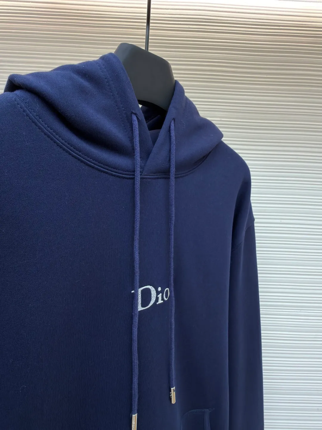 Dior classic logo embroidered hooded sweatshirt ●Item number: 1682333-A75 ●Material: cotton fabric ●Technology: embroidery production ●Color: navy blue ●Version: fitted version ●Weight: about 730 grams ●Size: XS-S-M-L ●Description: It adopts a fleece fabric structure of 26 counts of double yarn + 10S plush bottom, with a weight of 380g/㎡, and 21 counts of 1x1 ribs with a weight of 470g/㎡. It is used for the cuffs and hem. Its extremely high weight ensures the ultimate thickness, stiffness and deformation resistance of these closing parts, forming a unified and strong outline with the body. The fabric is a heavyweight and high-quality sweatshirt fabric. The 26-count double yarn forms a smooth, delicate and strong outer surface of the fabric, ensuring good color fastness and appearance. The 10S plush base undergoes a precise fleece or brushing process to form a layer of rich, dense and uniform fluff on the inside of the fabric. This makes the sweatshirt have a crisp and skeletal feel on the outside and an extremely soft and warm feel on the inside. White tone twill needle embroidery process. Needle embroidery is a straight line embroidery with stitches closely connected and evenly arranged. It is often used to outline clear and sharp outlines. This stitch is used to embroider a diagonal stripe pattern with a certain angle and width. Use white embroidery thread to follow the preset diagonal trajectory and perform precise embroidery with high-density stitches. On the high-weight fabric base, simple, exquisite and three-dimensional line decoration is created. The contrast between white and fabric color is sharp, and the slightly convex texture brought by the embroidery adds a visual and tactile layer, effectively enhancing the exquisite style of the basic sweatshirt. The overall wiring is straight and delicate, the fabric is comfortable and soft, and the color is consistent without color difference. It can be matched with the same style to create a fashionable look and control various occasions. Customized accessories, complete with three standards, same style for men and women!