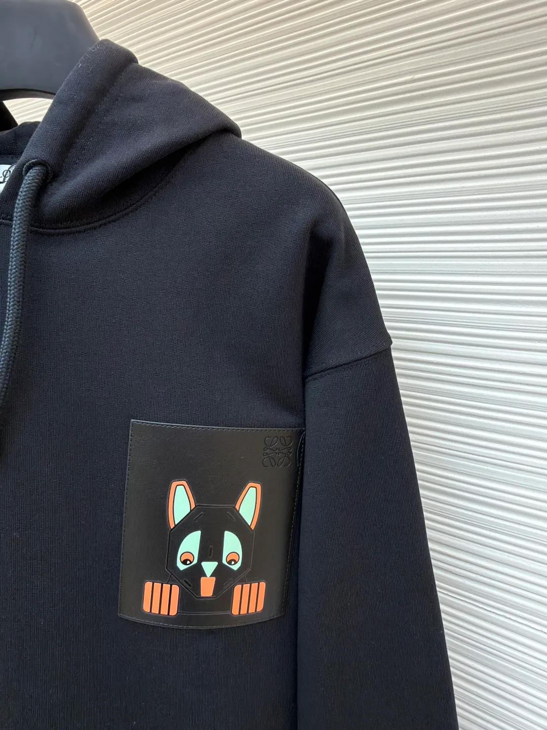 Loewe quirky kitten embossed hooded sweatshirt ●Original version: Purchased and developed by 8718 ●Item number: 1681954-A85 ●Material: pure cotton fabric ●Technology: genuine leather embossed production ●Color: black ●Version: fitted version ●Weight: about 920 grams ●Size: XS-S-M-L ●Description: This LV black hooded sweatshirt is made of 380g heavyweight fleece sweatshirt fabric. It is pre-shrunk and has a stable shape, with a fine matte texture on the surface. The hat rope is made of nylon braided rope + metal bell. The rope head is polished and rhodium-plated, with a cool texture and anti-oxidation. The chest patch pocket uses genuine leather embossing technology: the leather base is embossed with brand dark patterns, and the bag surface pattern uses multi-color silk screen printing + partial flocking. The ears, paws and other details of the animal image are raised by flocking to enhance the three-dimensional effect, and the color fastness reaches level 4 or above. The body stitching adopts double-needle overlocking technology, and the ribbing on the cuffs and hem is made of the same fabric, with an elastic rebound rate of ≥85%. The overall cutting error is controlled within 0.3cm, and the brand logo is embossed on the inside of the neckline, taking into account concealment and recognition, and balancing practicality and design. The overall wiring is straight and delicate, the fabric is comfortable and soft, and the color is consistent without color difference. It can be matched with the same style to create a fashionable look and control various occasions. Customized accessories, complete with three standards, same style for men and women!