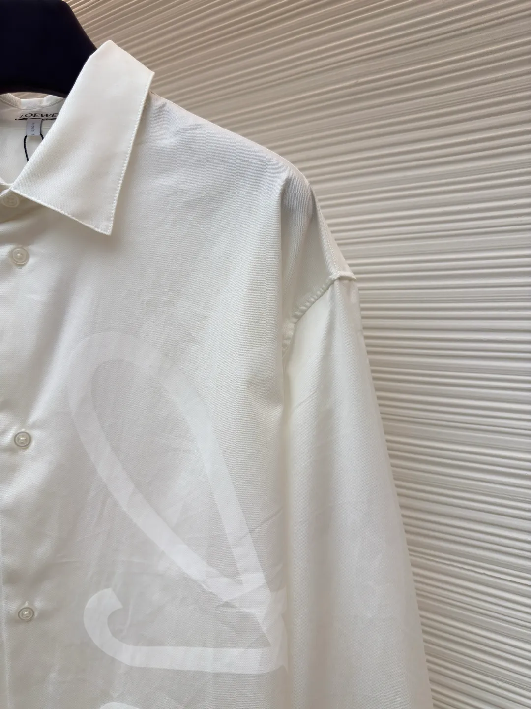 Loewe concealed logo lapel shirt ●Original version: Purchased and developed by 5268 ●Item No.: 1680722-A75 ●Fabric: Light cotton poplin shirt fabric ●Technology: Printing ●Version: Fitted version ●Weight: about 270 grams ●Size: 36-38-40-42 ●Description: Poplin is a classic cotton fabric characterized by a large density difference between warp and weft yarns, forming a fine diamond-shaped grainy texture. This structure not only gives the fabric a refreshing and breathable wearing feel, but also ensures that its texture is light and thin but has a crisp texture, avoiding the shortcomings of ordinary thin cotton that easily collapses, perfectly supporting the neat silhouette of the shirt, while maintaining the unique skin-friendly comfort of cotton. The chest is decorated with a discharge printing process; discharge printing is a special process that erodes through the dyed base fabric to reveal the light-colored pattern of the original fabric. This makes the Anagram logo not simply covered on the fabric, but integrated with the fabric, feeling smooth and traceless, showing a low-key and high-end matte effect. Fit and design The shirt adopts a loose fit of standard length; it is precisely tailored to the pattern. It is neither too loose nor deliberately slim, aiming to create a clean silhouette and is extremely tolerant to all body types. The standard length ensures that the hem can be tucked into trousers or hang naturally, meeting a variety of wear needs, including formal and casual. The overall wiring is straight and delicate, the fabric is comfortable and soft, and the color is consistent without color difference. It can be matched with the same style to create a fashionable look and control various occasions. Customized accessories, complete with three standards, same style for men and women!