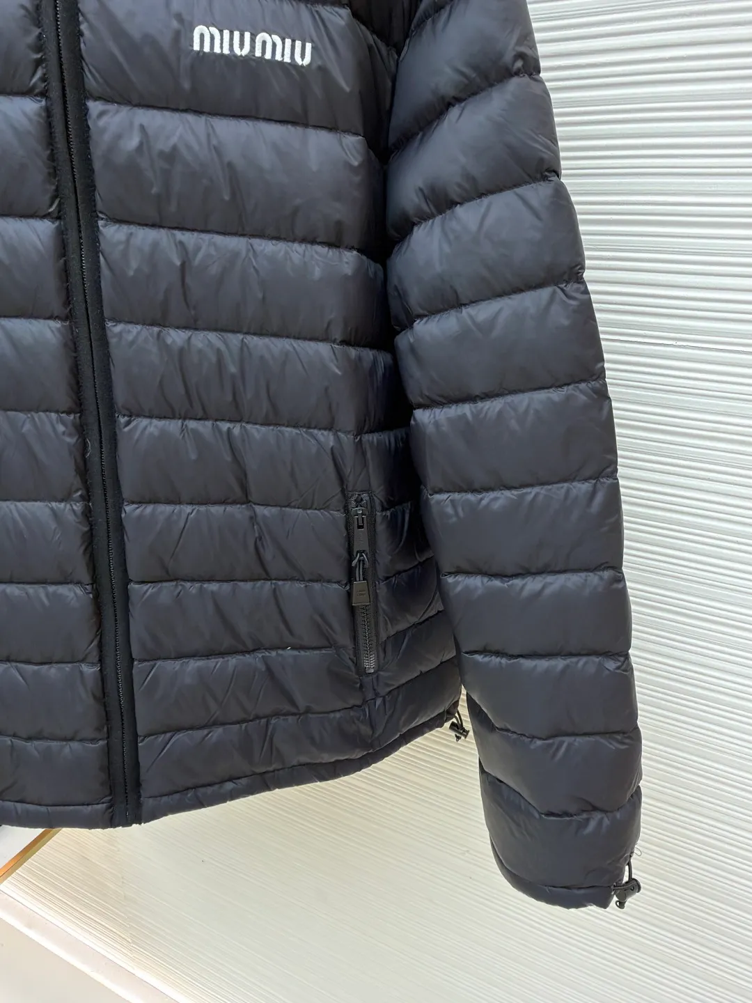 MIU MIU hooded down jacket ●Item number: 1681314-C55 ●Material: white duck down fabric ●Technology: mechanical down filling ●Color: black ●Version: fitted version ●Weight: about 615 grams ●Size: S-M-L-XL ●Description: This is a light down jacket from the MiuMiu brand. Its craftsmanship details are as follows: It is made of exquisite pongé fabric made of 100% recycled polyester fiber. This fabric has good windproof performance, and at the same time has a delicate texture and a high-end luster. It has high fluffiness, excellent warmth retention and is very light overall, allowing the wearer to gain warmth without any heavy burden. It has a three-dimensional cut and a neat silhouette. Precision stitching technology is used during sewing to form regular quilting lines, which not only serves to fix the down, but also enhances the structure and design of the garment. The stitches are fine and even, and the seams are smooth, reflecting the exquisite craftsmanship of garment making. Equipped with a customized wax zipper, the zipper is smooth and durable, and the zipper puller has brand details and a high-quality feel. The hooded design fits the head and increases warmth; the drawstring cuffs can be adjusted according to needs to prevent cold wind from entering; the elastic drawstring at the hem can adjust the looseness of the hem, improving the comfort and practicality of wearing; there is the brand's embroidered logo on the chest, with fine craftsmanship and highlighting the brand identity.