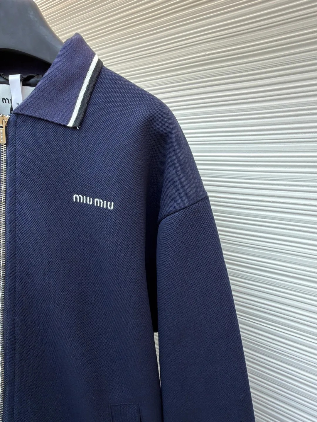 MIUMIU letter embroidered pique lapel jacket ●Original version: 25400 purchased and developed ●Item number: 1681338-B20 ●Material: combed cotton fabric ●Technology: embroidery production ●Color: navy ●Version: Fitted version ●Weight: about 1130 grams ●Size: 36-38-40-42 ●Description: This jacket is made of high-count combed cotton blended fabric, which is crisp and soft to the touch. The neckline is a classic lapel design, and the edges are decorated with double topstitching in contrasting colors. The black and white lines are precisely aligned, demonstrating the brand's control of details. The placket is equipped with a custom metal zipper. Its smoothness has been tested many times for wear resistance, and there is no lag when closing. The "miu miu" logo on the chest adopts high-precision embroidery technology, with fine and even stitching and full three-dimensionality. There are three-dimensional pockets on both sides of the body, and the edges of the pockets are hemmed to prevent the fabric from coming off. The ribs on the cuffs and hem are made of high-elastic knitting technology to ensure fit and resilience when worn. The overall tailoring follows an oversize silhouette, and the splicing of the body uses double stitching + overlocking techniques to ensure fastness and prevent threads from being exposed. Every craft detail reflects the brand's high-end clothing making standards.