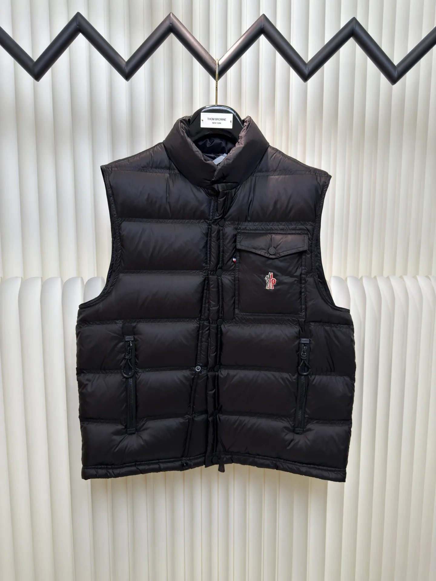 P700 MONCLER masked down vest with reflective markings is made of 90 white duck down with a weight of 130g to ensure excellent thermal insulation performance. After strict screening and processing, the velvet is not easy to get drilled, ensuring durability.  It is made of extremely fine nylon yarn and is woven in a high-density manner. The number of yarns per unit area is large, and the gaps between the yarns are very small, which can effectively block the penetration of cold wind. The fine weave keeps it lightweight and soft to the touch, physically restricting the lightweight down fibers from slipping through the gaps. Nylon fiber itself has good wear resistance and is not easy to snag, which significantly extends its service life and can easily cope with the needs of multiple scenarios.  The lining is made of high-density weaving of 100% polyester fiber, with a thickness of 0.08mm to avoid increasing the overall weight and making it smooth to wear. The high-density weaving structure forms a wind resistance layer to protect the thermal efficiency of the thermal layer from loss. It can withstand the expansion pressure of down and prevent deformation and damage after long-term use.  Through the reflective cloth punching process, the logo is regularly arranged on the triangular reflective mark with dozens of small holes, which not only improves functionality but also enhances recognition and aesthetics. Size 1 2 3 4