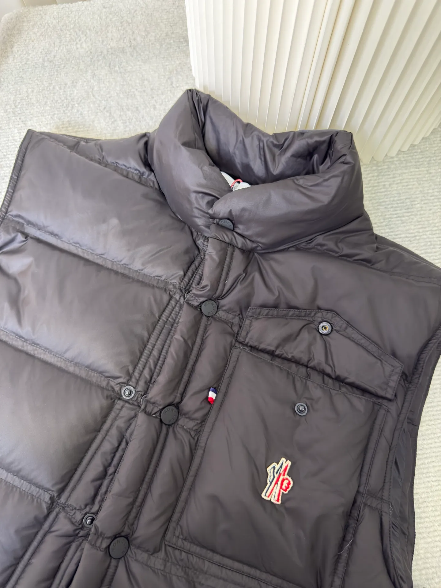 P700 MONCLER masked down vest with reflective markings is made of 90 white duck down with a weight of 130g to ensure excellent thermal insulation performance. After strict screening and processing, the velvet is not easy to get drilled, ensuring durability.  It is made of extremely fine nylon yarn and is woven in a high-density manner. The number of yarns per unit area is large, and the gaps between the yarns are very small, which can effectively block the penetration of cold wind. The fine weave keeps it lightweight and soft to the touch, physically restricting the lightweight down fibers from slipping through the gaps. Nylon fiber itself has good wear resistance and is not easy to snag, which significantly extends its service life and can easily cope with the needs of multiple scenarios.  The lining is made of high-density weaving of 100% polyester fiber, with a thickness of 0.08mm to avoid increasing the overall weight and making it smooth to wear. The high-density weaving structure forms a wind resistance layer to protect the thermal efficiency of the thermal layer from loss. It can withstand the expansion pressure of down and prevent deformation and damage after long-term use.  Through the reflective cloth punching process, the logo is regularly arranged on the triangular reflective mark with dozens of small holes, which not only improves functionality but also enhances recognition and aesthetics. Size 1 2 3 4