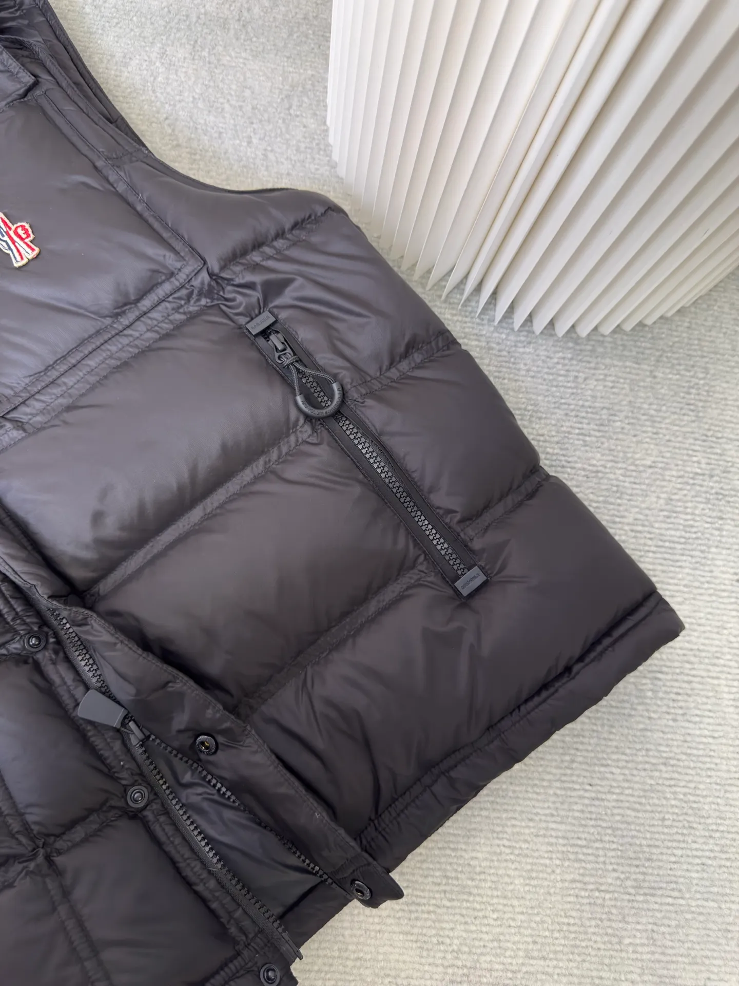 P700 MONCLER masked down vest with reflective markings is made of 90 white duck down with a weight of 130g to ensure excellent thermal insulation performance. After strict screening and processing, the velvet is not easy to get drilled, ensuring durability.  It is made of extremely fine nylon yarn and is woven in a high-density manner. The number of yarns per unit area is large, and the gaps between the yarns are very small, which can effectively block the penetration of cold wind. The fine weave keeps it lightweight and soft to the touch, physically restricting the lightweight down fibers from slipping through the gaps. Nylon fiber itself has good wear resistance and is not easy to snag, which significantly extends its service life and can easily cope with the needs of multiple scenarios.  The lining is made of high-density weaving of 100% polyester fiber, with a thickness of 0.08mm to avoid increasing the overall weight and making it smooth to wear. The high-density weaving structure forms a wind resistance layer to protect the thermal efficiency of the thermal layer from loss. It can withstand the expansion pressure of down and prevent deformation and damage after long-term use.  Through the reflective cloth punching process, the logo is regularly arranged on the triangular reflective mark with dozens of small holes, which not only improves functionality but also enhances recognition and aesthetics. Size 1 2 3 4