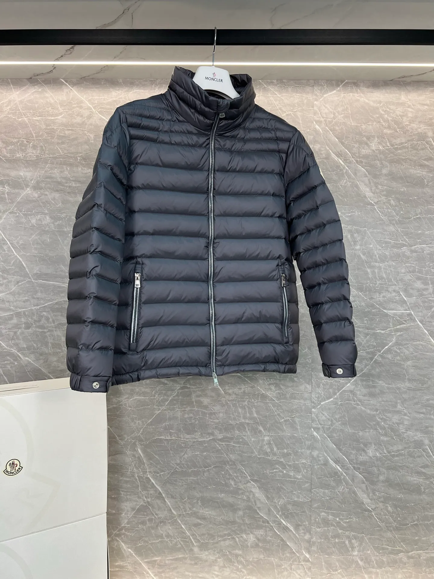 P780 Moncler Arnique short and lightweight men's down jacket. Super soft and delicate fabric, white duck down filling. Extremely lightweight and warm. The Arnique hooded down jacket is made of polyester fiber outer fabric and is Moncler's classic men's design item.  The Arnique short down jacket features classic checkered quilting and signature patch pockets on the sleeves, highlighting the Moncler style. The outer fabric reflects the brand's commitment to lowering its environmental footprint. Modular design, removable hood can be easily put on and taken off according to weather conditions. Details Made of polyester outer fabric Polyester lining Filled with down Size: 1-5 Silver Gray Black