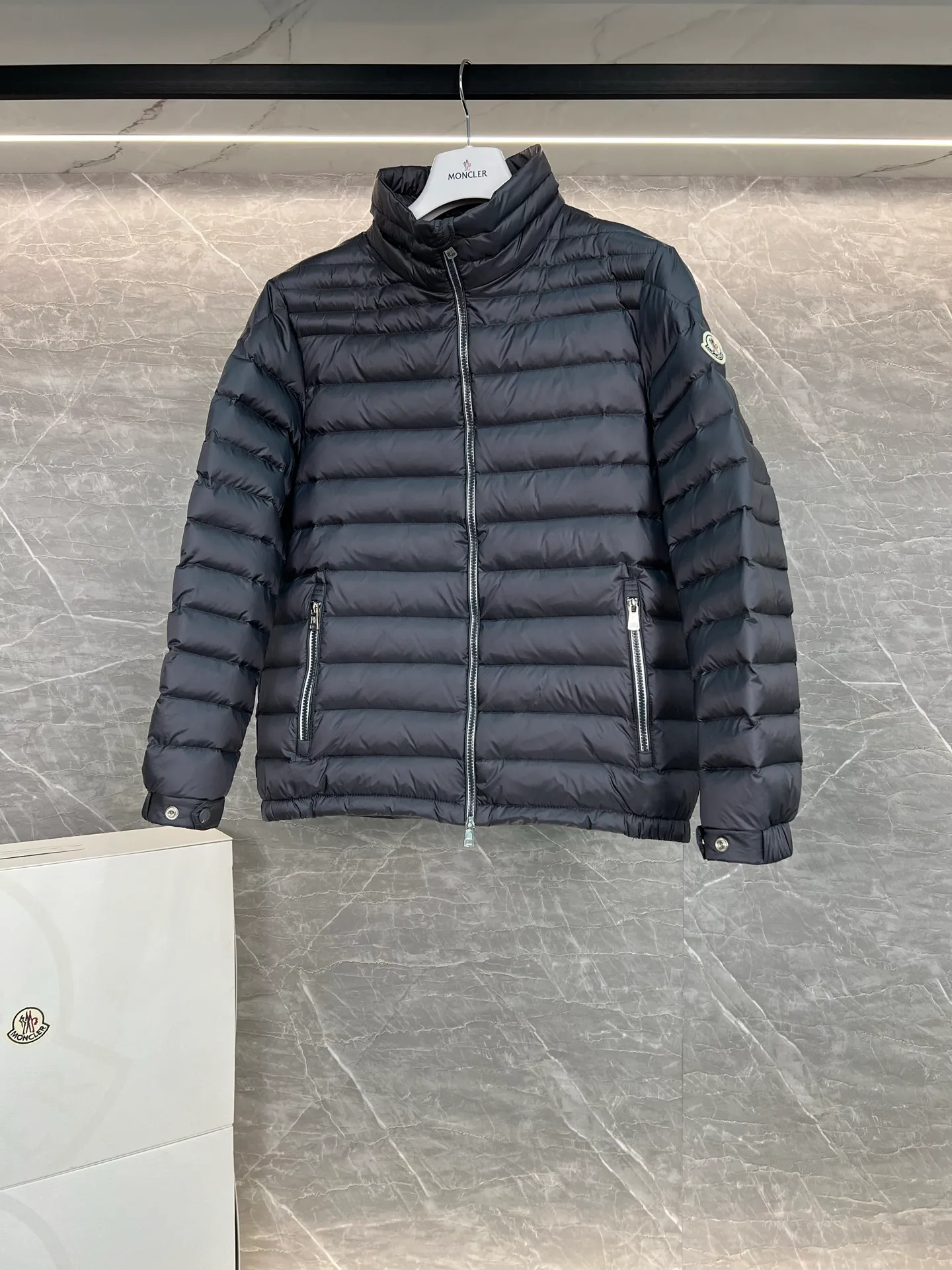 P780 Moncler Arnique short and lightweight men's down jacket. Super soft and delicate fabric, white duck down filling. Extremely lightweight and warm. The Arnique hooded down jacket is made of polyester fiber outer fabric and is Moncler's classic men's design item.  The Arnique short down jacket features classic checkered quilting and signature patch pockets on the sleeves, highlighting the Moncler style. The outer fabric reflects the brand's commitment to lowering its environmental footprint. Modular design, removable hood can be easily put on and taken off according to weather conditions. Details Made of polyester outer fabric Polyester lining Filled with down Size: 1-5 Silver Gray Black