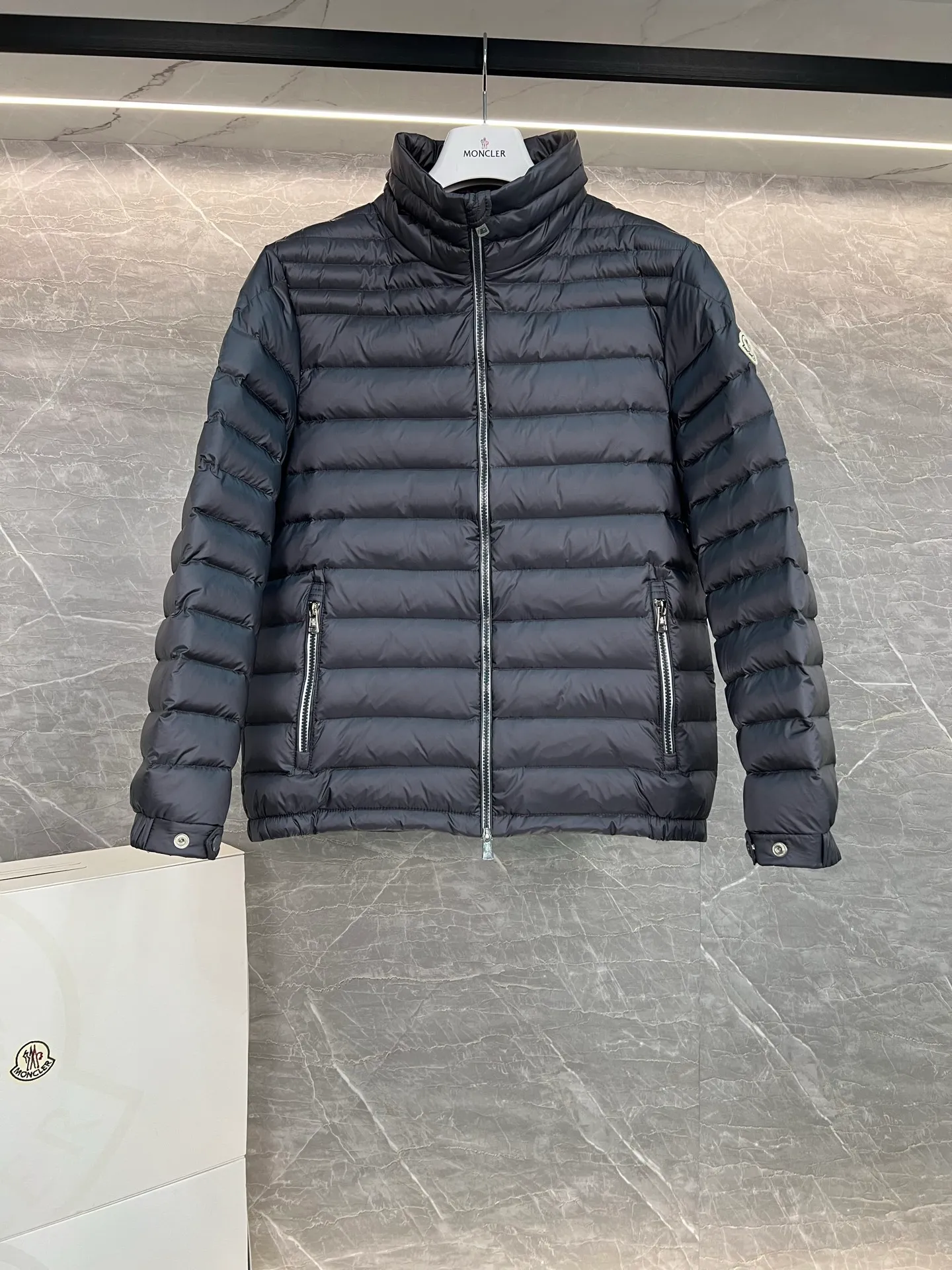 P780 Moncler Arnique short and lightweight men's down jacket. Super soft and delicate fabric, white duck down filling. Extremely lightweight and warm. The Arnique hooded down jacket is made of polyester fiber outer fabric and is Moncler's classic men's design item.  The Arnique short down jacket features classic checkered quilting and signature patch pockets on the sleeves, highlighting the Moncler style. The outer fabric reflects the brand's commitment to lowering its environmental footprint. Modular design, removable hood can be easily put on and taken off according to weather conditions. Details Made of polyester outer fabric Polyester lining Filled with down Size: 1-5 Silver Gray Black