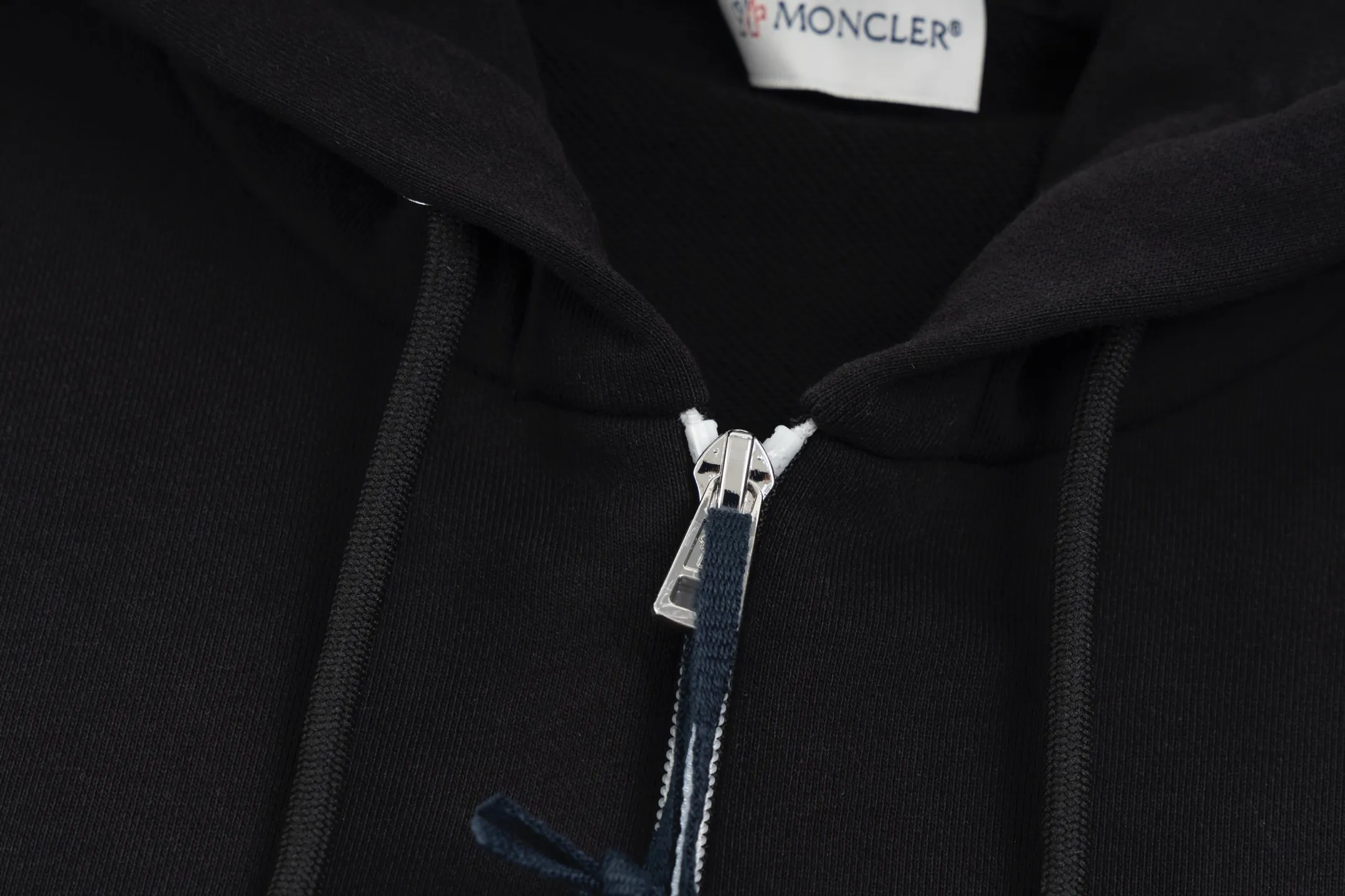 P480 Exclusive supply] Moncler slogan printed zipper hooded jacket is made of lightweight turned velvet cotton fabric. High-density cotton fiber is used to achieve internal and external differentiation in a single layer of fabric. The outer plain weave is tight and wear-resistant, and the inner layer is formed into a short dense suede surface through a sanding process. The dense plain weave structure resists wind and splashes, the brushed suede surface is skin-friendly, and the short pile has been treated to eliminate static electricity to avoid sticking to the skin. A very small amount of grooved polyester moisture-conducting yarn is embedded in the cotton fiber, which quickly guides sweat from the inner suede surface to the outer layer for evaporation through the capillary effect, solving the problem of the suede being easily stuffy. By actively interfering with the shrinkage potential of cotton fibers, the fabric is able to transition from changeable to stable, improving dimensional stability while being less prone to deformation and wrinkles. A high-saturation pattern is displayed on the center of the cap through glue printing. The scraper pressure is even and the scraping is performed in 5 steps to ensure that the glue is fully penetrated and covered. After curing at high temperature, the pigment is firmly bonded to the cotton fiber. The ink-absorbing and penetrating properties of the cotton fiber make the pattern less likely to fall off due to friction. Size:S/M/L/XL