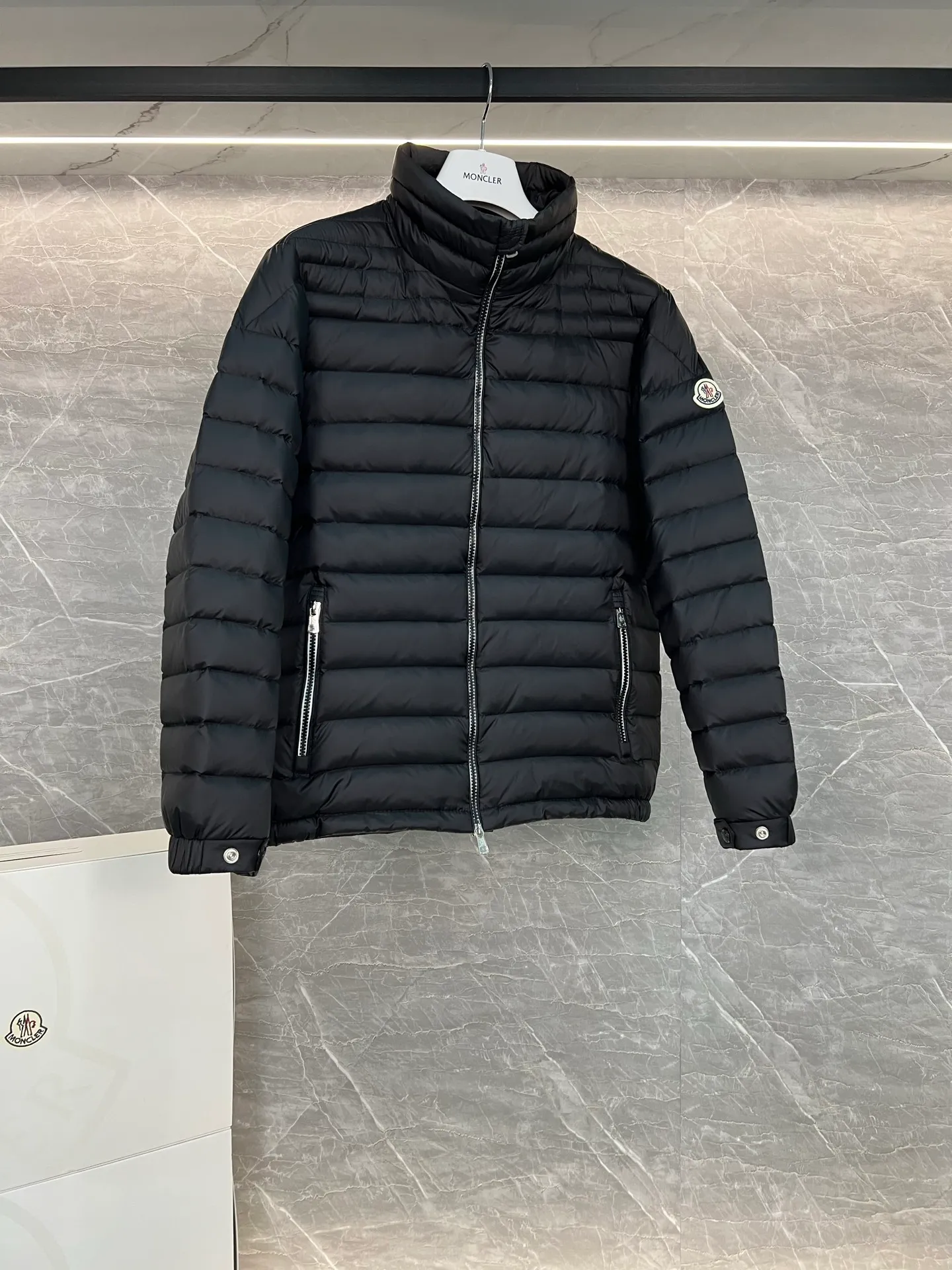 P780 Moncler Arnique short and lightweight men's down jacket. Super soft and delicate fabric, white duck down filling. Extremely lightweight and warm. The Arnique hooded down jacket is made of polyester fiber outer fabric and is Moncler's classic men's design item.  The Arnique short down jacket features classic checkered quilting and signature patch pockets on the sleeves, highlighting the Moncler style. The outer fabric reflects the brand's commitment to lowering its environmental footprint. Modular design, removable hood can be easily put on and taken off according to weather conditions. Details Made of polyester outer fabric Polyester lining Filled with down Size: 1-5 Silver Gray Black