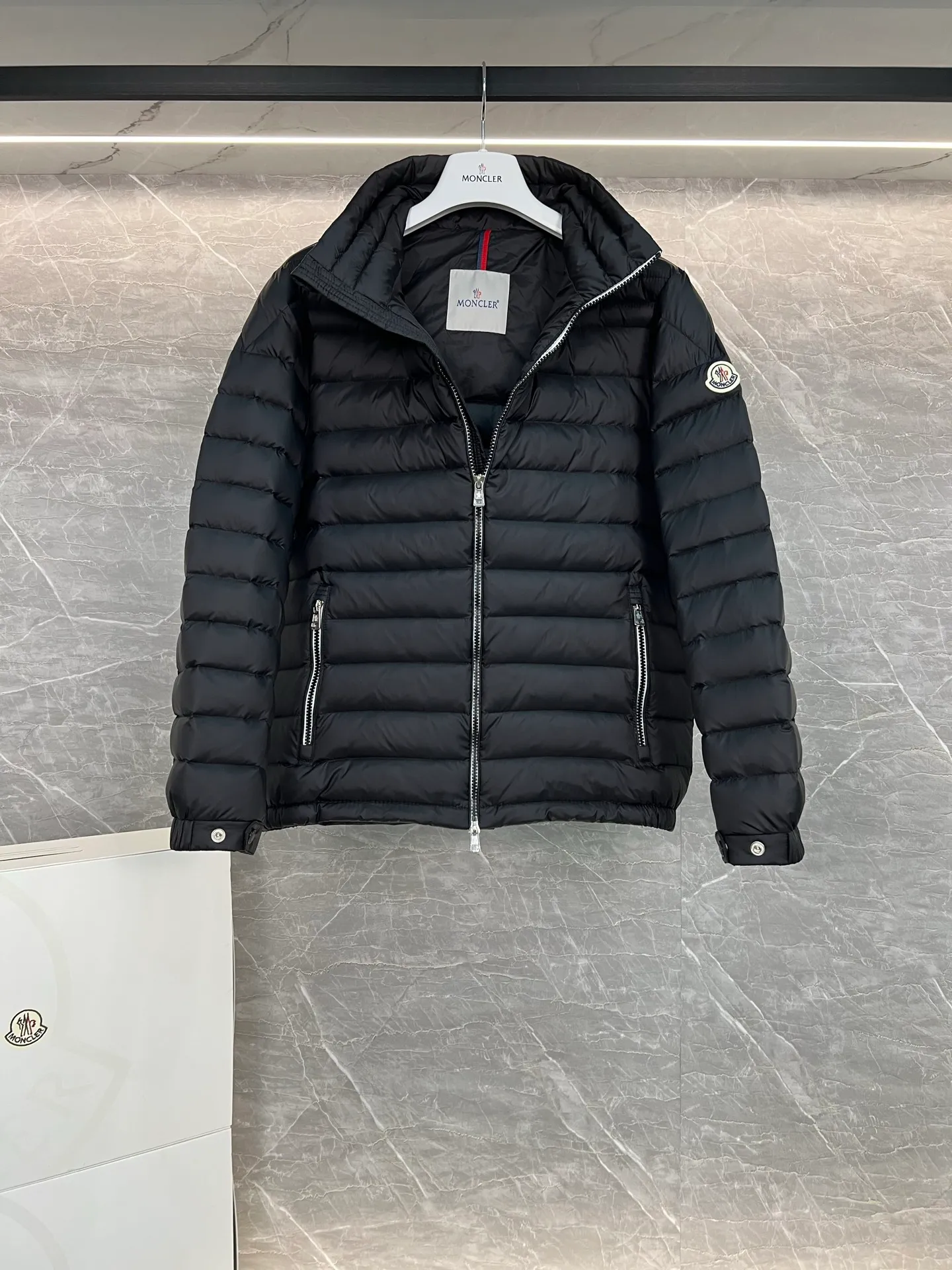 P780 Moncler Arnique short and lightweight men's down jacket. Super soft and delicate fabric, white duck down filling. Extremely lightweight and warm. The Arnique hooded down jacket is made of polyester fiber outer fabric and is Moncler's classic men's design item.  The Arnique short down jacket features classic checkered quilting and signature patch pockets on the sleeves, highlighting the Moncler style. The outer fabric reflects the brand's commitment to lowering its environmental footprint. Modular design, removable hood can be easily put on and taken off according to weather conditions. Details Made of polyester outer fabric Polyester lining Filled with down Size: 1-5 Silver Gray Black