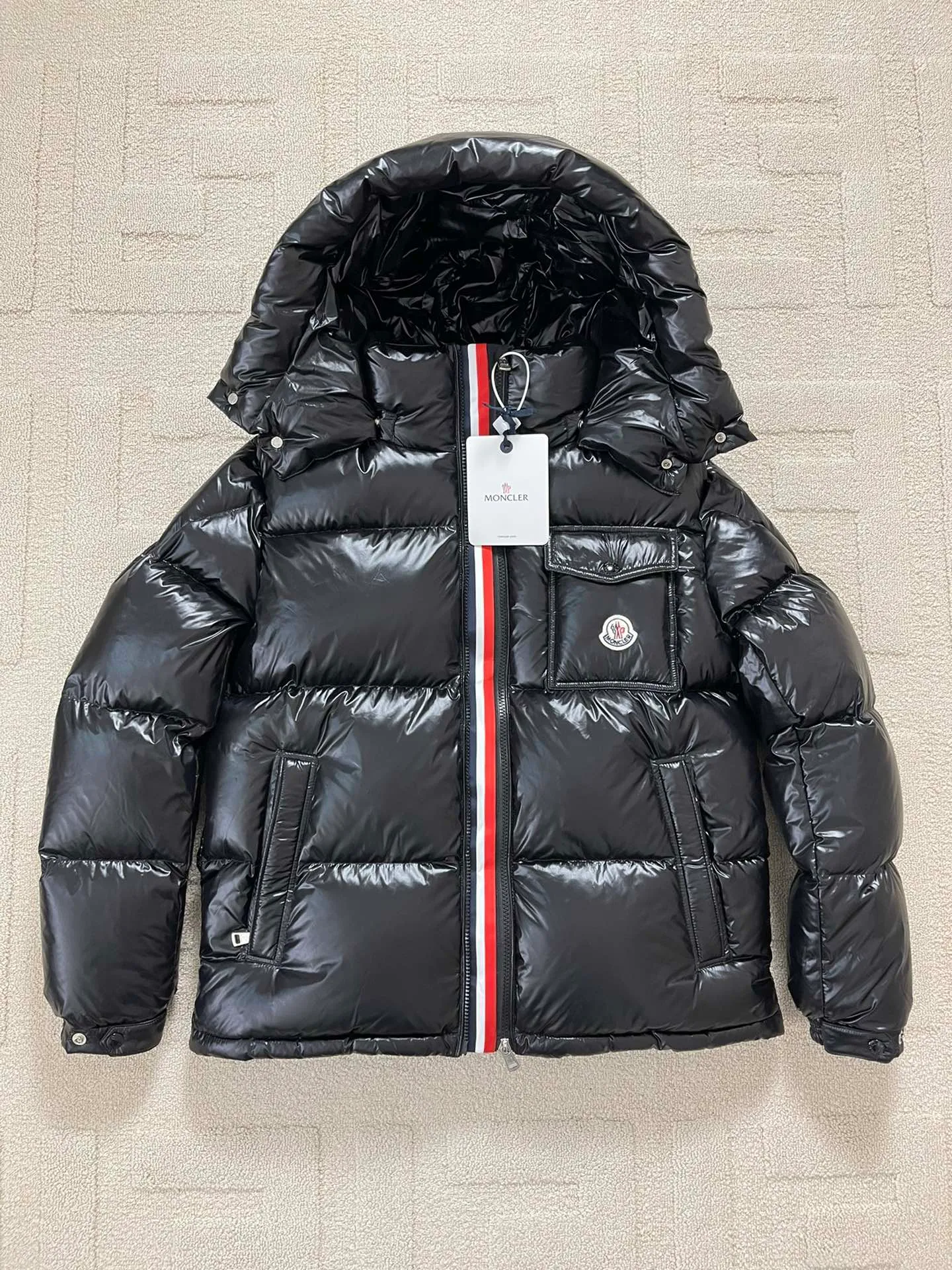 Flat detail P1180 Moncler Montbeliard glossy down jacket The Montbeliard down jacket is made of shiny patented nylon and features highly recognizable rectangular quilting. Featuring a hooded design and signature patch pocket at chest.  Fabric: 100% nylon Filling content: 90% white goose down 250-300g Craftsmanship: Made of nylon laqué fabric Nylon laque lining Filled with down, detachable and adjustable hood with drawstring closure Built-in flap with webbing, black pocket details at the lower end Zipper closure Collar with snap buttons Zipper pockets Patch pockets with snap buttons Adjustable cuffs Adjustable elastic drawstring hem.  Size: 1 2 3 4 5 0548000