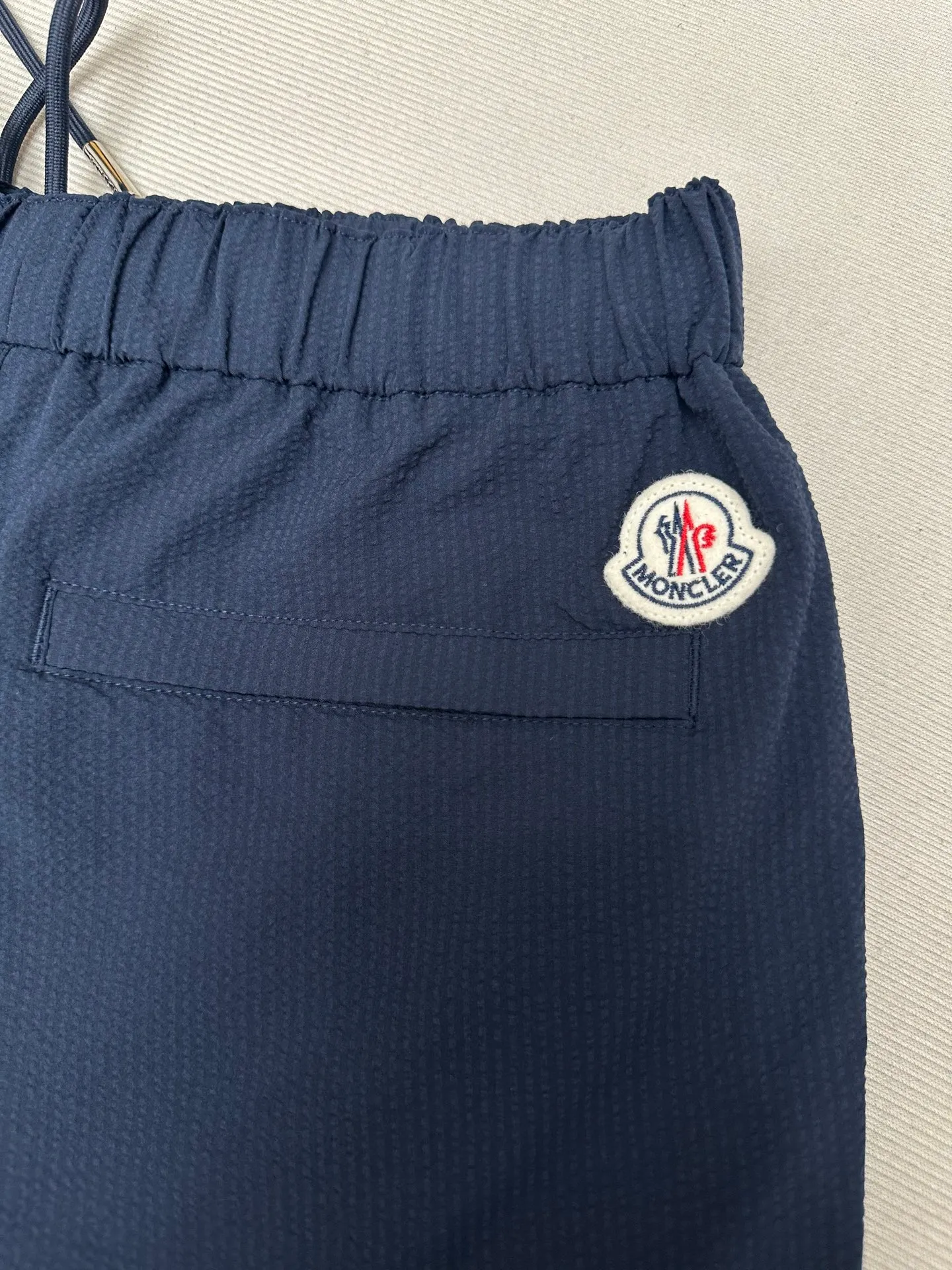 P320 MONCLER Puff Wrinkle Logo Shorts Fabric Selection: Made of cotton blended woven fabric with 76% high cotton content + 24% polyester fiber. The high cotton content ensures that the fabric is soft, skin-friendly, and has good breathability. The addition of a small amount of polyester fiber balances the wrinkle resistance and elasticity of the overall fabric and greatly reduces its shrinkage. After preshrinking, dimensional changes during washing are negligible. The fusion of the two makes the fabric combine anti-wrinkle, quick-drying, easy care and other characteristics without losing the advantages of good wearing experience and strong breathability.  Bubble wrinkle technology: ground warp + bubble warp independent let-off. The bubble warp yarn feeding speed is about 20% higher than the ground warp yarn feeding speed. During weaving, the bubble warp is relaxed and accumulated, forming a naturally arched bubble unit. Through the alternating arrangement of yarns + floating length tissue changes, a clear grid frame is built between the bubble units, and finally the bubble three-dimensional effect and grid outline are locked through high-temperature heat setting.  Accessories: One-to-one customized drawstring head One-to-one customized LOGO labeling SIZE: 1 2 3 4