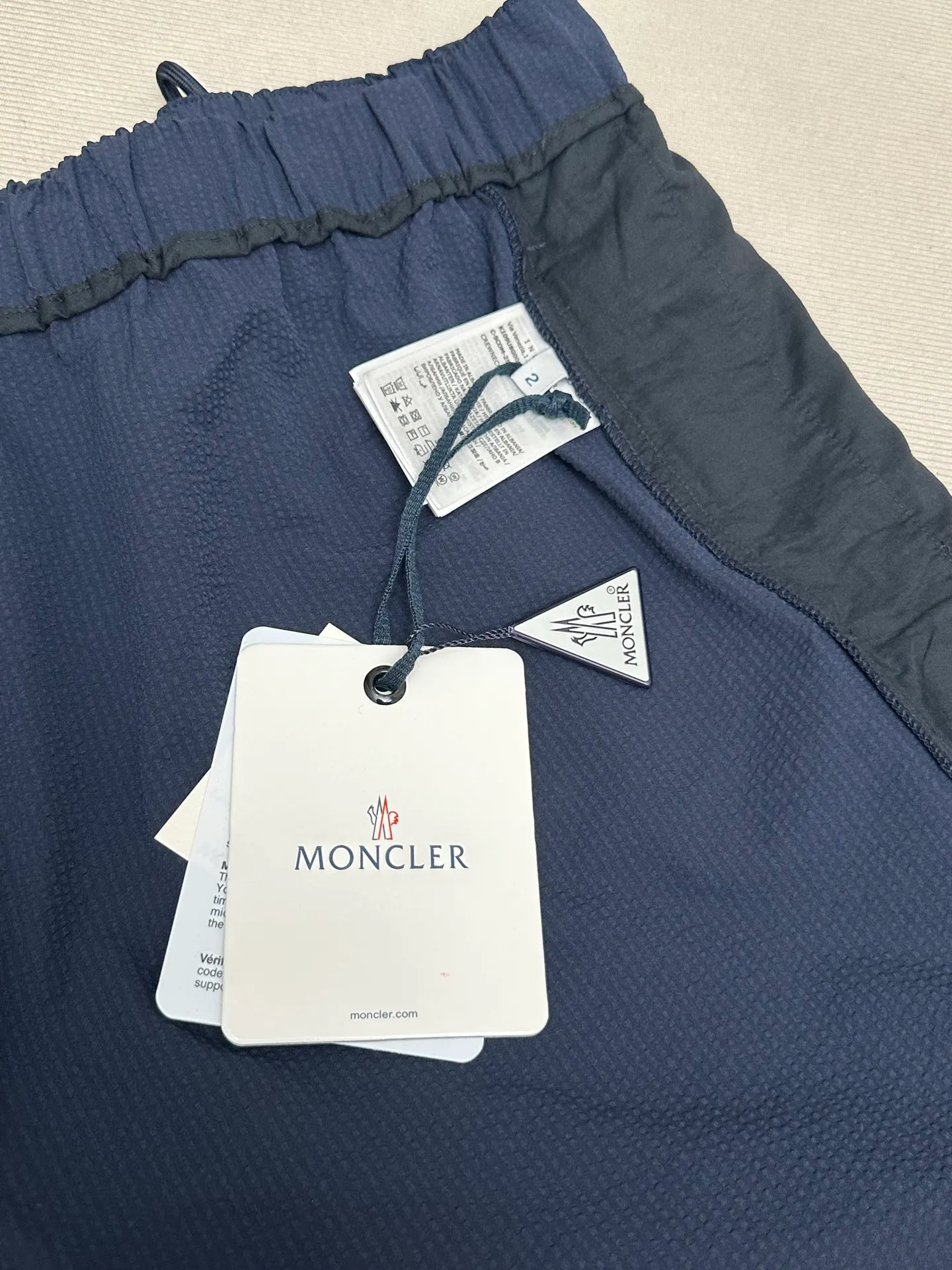 P320 MONCLER Puff Wrinkle Logo Shorts Fabric Selection: Made of cotton blended woven fabric with 76% high cotton content + 24% polyester fiber. The high cotton content ensures that the fabric is soft, skin-friendly, and has good breathability. The addition of a small amount of polyester fiber balances the wrinkle resistance and elasticity of the overall fabric and greatly reduces its shrinkage. After preshrinking, dimensional changes during washing are negligible. The fusion of the two makes the fabric combine anti-wrinkle, quick-drying, easy care and other characteristics without losing the advantages of good wearing experience and strong breathability.  Bubble wrinkle technology: ground warp + bubble warp independent let-off. The bubble warp yarn feeding speed is about 20% higher than the ground warp yarn feeding speed. During weaving, the bubble warp is relaxed and accumulated, forming a naturally arched bubble unit. Through the alternating arrangement of yarns + floating length tissue changes, a clear grid frame is built between the bubble units, and finally the bubble three-dimensional effect and grid outline are locked through high-temperature heat setting.  Accessories: One-to-one customized drawstring head One-to-one customized LOGO labeling SIZE: 1 2 3 4