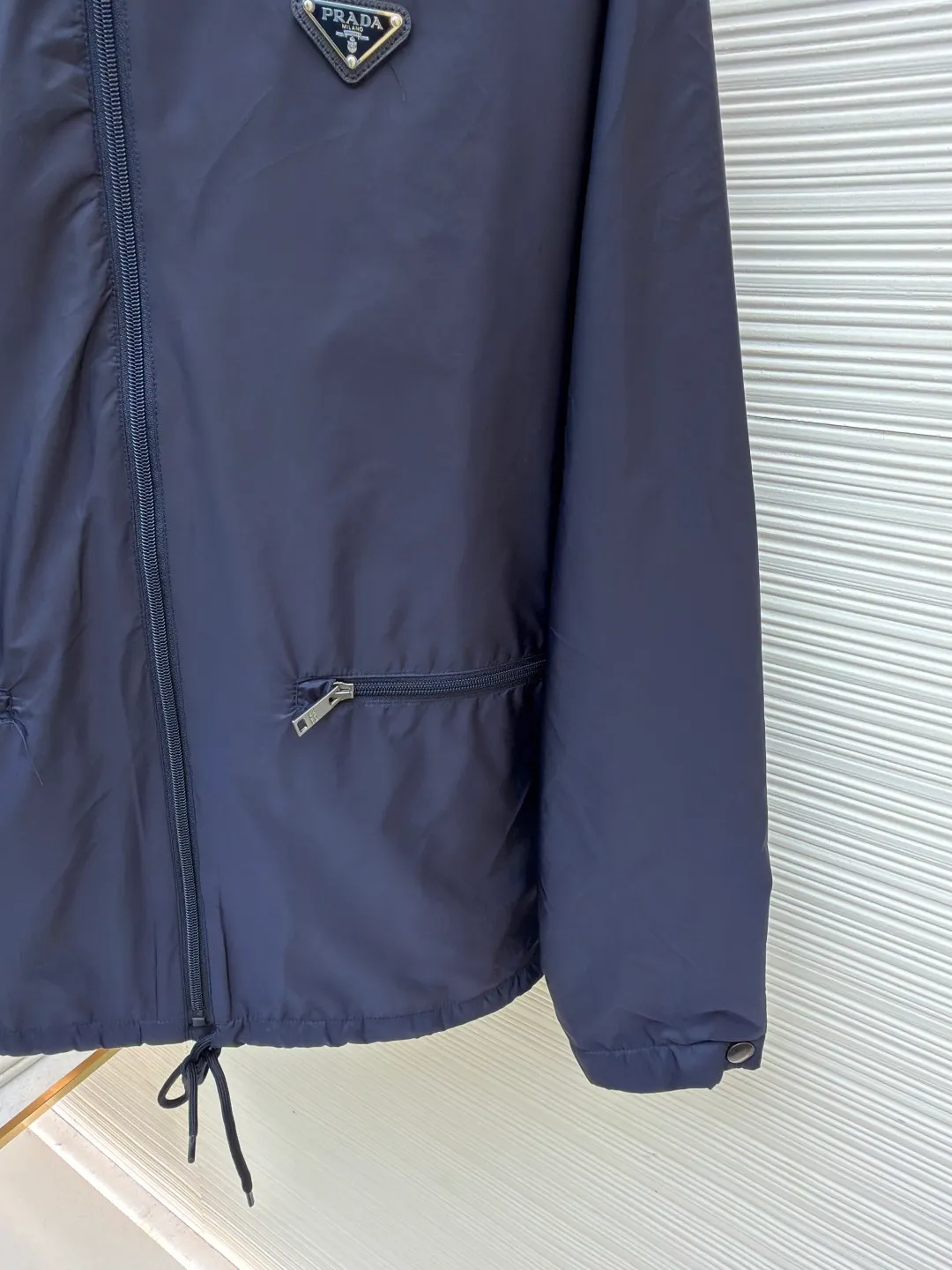 Prada Regenerated Nylon Fleece Jacket ●Original version: Purchased and developed by 12048 ●Item number: 1682331-B70 ●Fabric: Regenerated nylon fabric ●Technology: Made on positioning cutting machine ●Color: Navy blue ●Version: Fitted version ●Weight: about 585 grams ●Size: S-M-L-XL ●Description: High-density yarn is made by melt spinning through a special process. The yarn has been stretched and twisted through multiple processes to give the fabric the stiffness and wrinkle resistance that are comparable to those of native materials, while retaining a light and drapey wearing experience. The surface of the fabric is treated with an environmentally friendly coating process to form a micro-waterproof film layer, which is both windproof and breathable. The main raw material is polyester fiber blended wool raw material which is cleaned and pre-treated to remove impurities. After the yarn is woven into fabric through a loom, it is brushed, that is, the surface of the fiber is fluffed through mechanical brushing to form fluff. Then the shaking process is carried out, and the fabric is shaken under high temperature and humid conditions, so that the fibers form small spherical particles to increase the thermal insulation performance. The shoulder splicing uses twill sewing technology, the raglan sleeve design releases shoulder movement space, and the regular shoulder line ensures the stiffness of the overall silhouette, maintaining an elegant silhouette for both sports and static wear. The three-dimensional pleat design is incorporated into both sides of the body and sleeve elbows, and a long-lasting pleat texture is formed through high-temperature setting technology. This not only enhances the aerodynamic performance of the fabric, but also enriches the visual hierarchy with light and shadow changes, breaking the dull feeling of solid color fabrics. The cuffs are equipped with a hidden snap-button adjustment system, which can be freely tightened according to the circumference of the wrist to prevent cold wind from entering; the straight bottom edge is matched with a drawstring at the hem, and the inner rope buckle design can be used to adjust the tightness, taking into account warmth preservation and waistline shaping. The overall wiring is straight and delicate, the fabric is comfortable and soft, and the color is consistent without color difference. It can be matched with the same style to create a fashionable look and control various occasions. Customized accessories, complete with three standards, same style for men and women!